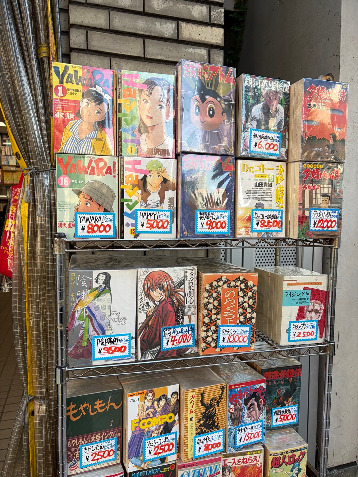 Manga sets including YAWARA!, HAPPY!, and Astro Boy displayed on metal shelves with handwritten price tags.